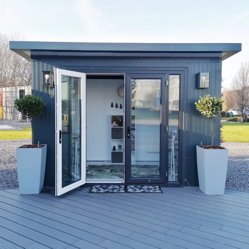 Render showing blue-grey garden room with large glass sliding doors, modern interior with shelving and seating area, wooden decking, landscaping with plants in pots