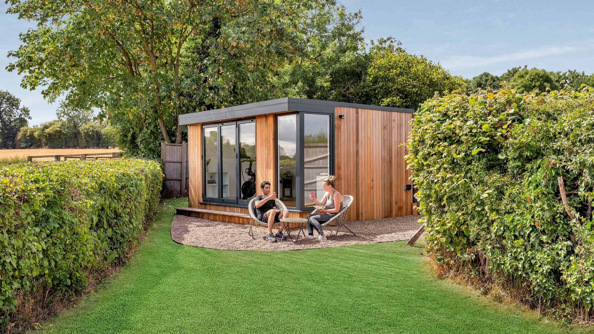 Exterior garden room with wooden cladding, large windows, two people sitting outside on patio, surrounded by green hedges and trees